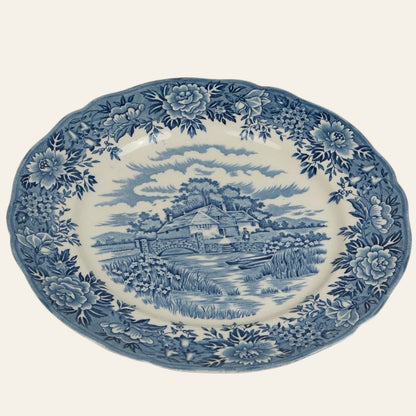 A vintage white dinner plate with a classic blue and white transferware design depicting a colonial village scene, including a thatched-roof cottage and a stone bridge.