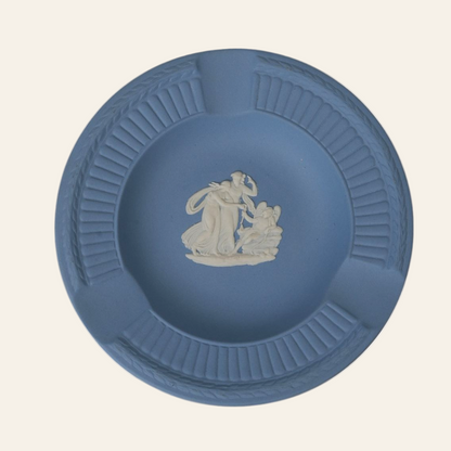 A small, round, matte blue Wedgwood Jasperware plate with a white relief figure of a classical scene in the center and laurel garland detailing along the rim.
