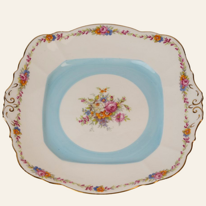 A vintage blue floral cake plate with gold trim and a scalloped edge, featuring a bouquet of flowers in the center.