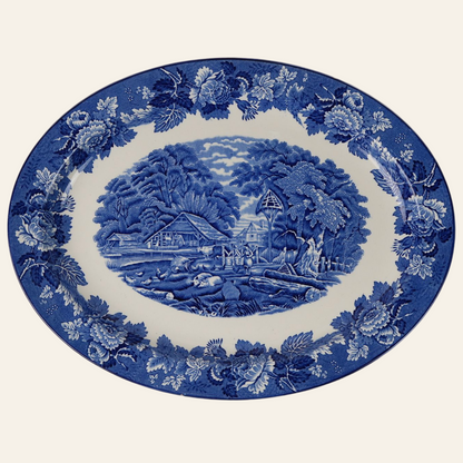 A blue and white oval ceramic platter with an intricate pastoral scene in the center and a floral border.