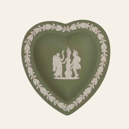 A small heart-shaped ceramic trinket plate with a white relief scene of two figures on it, bordered by a laurel wreath.