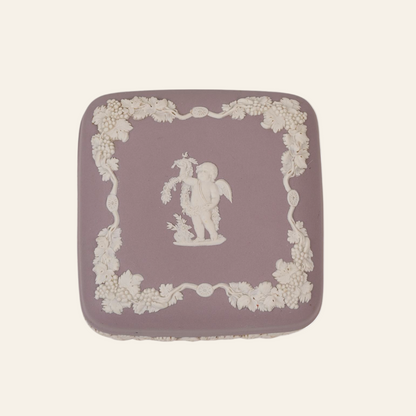 A lilac-colored vintage Wedgwood trinket box with a floral pattern and a figure in the center, framed by a white border.