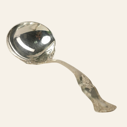 An antique silver-plated ladle with an ornately designed handle featuring grapevine motifs and a scalloped, deep bowl with embossed scrollwork along the edges.