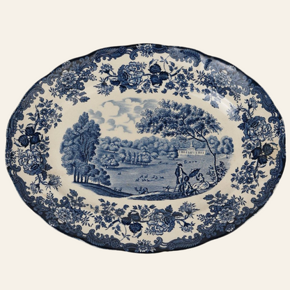 A round, blue and white hand-engraved ceramic platter with a floral border and a central scene featuring trees, a man, a woman, and a dog.
