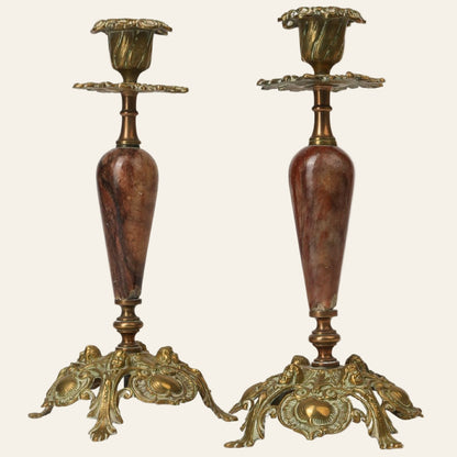 Two antique brass and marble candle stands with ornate bases and floral-inspired designs on the brass columns.