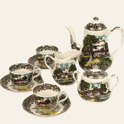 A vintage coffee service set featuring a floral and rural scene pattern on a ceramic surface, including a teapot, sugar bowl, creamer, and teacups with saucers.