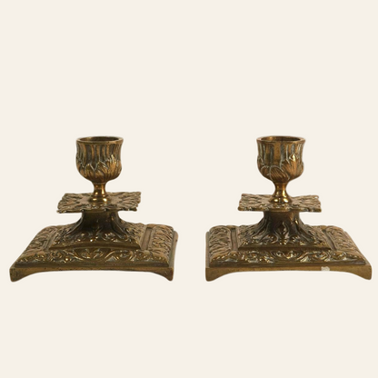 A pair of vintage brass candle holders with baroque-inspired details, including acanthus leaf patterns and a square base.