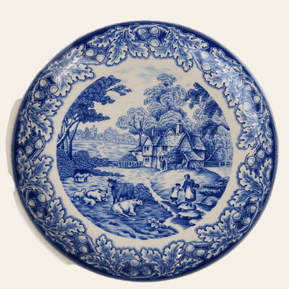 A round blue and white porcelain dinner plate with a floral border and a pastoral scene in the center, featuring cottages, trees, and figures.