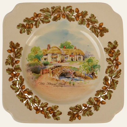 A small, beige Royal Doulton ceramic side plate with a hand-painted scene of an old English inn, featuring a thatched roof, a stone bridge, and greenery. The plate has a floral border with acorns and oak leaves.