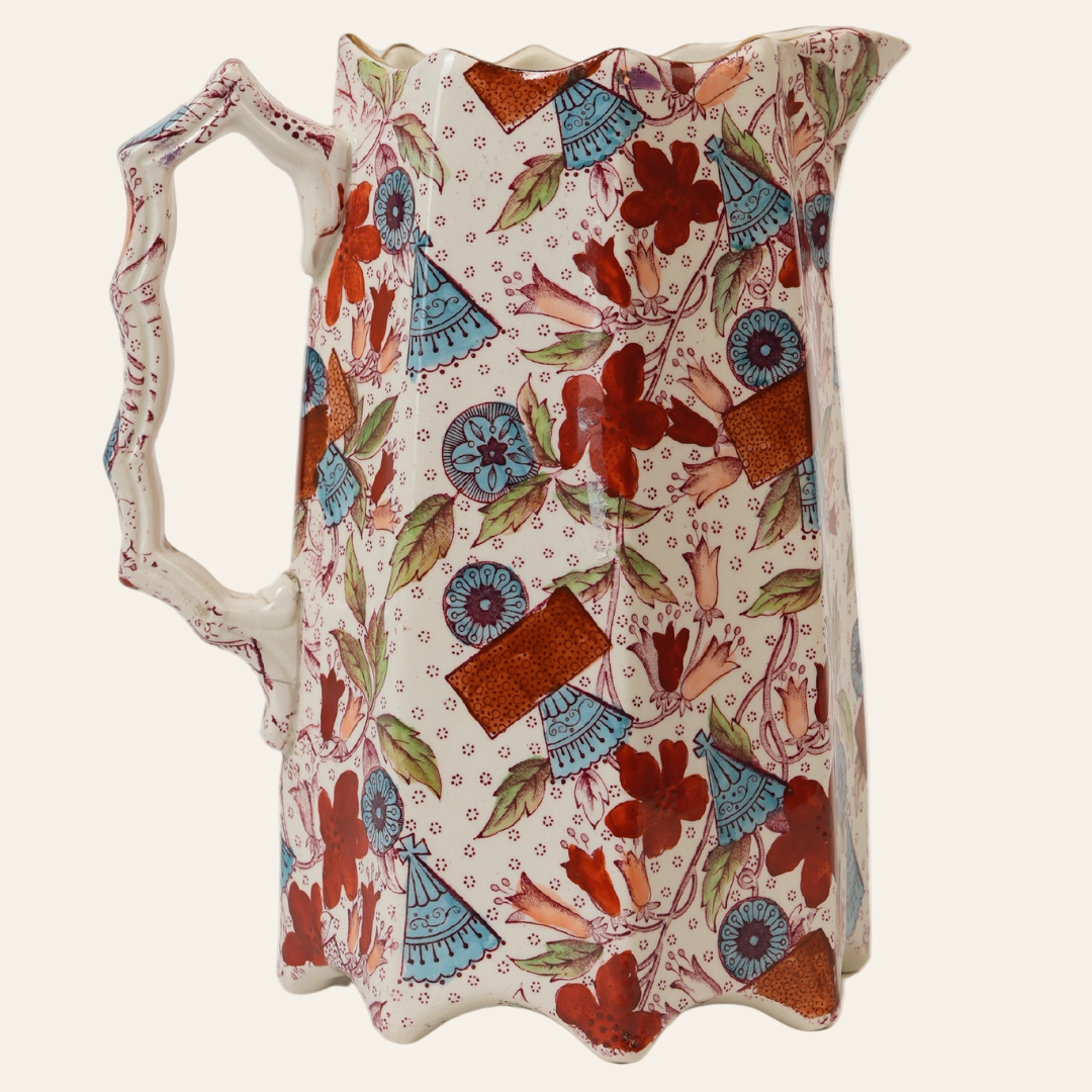 A vintage Imari-style porcelain pitcher with hand-painted florals, geometric medallions, and fan motifs in rust red, cobalt blue, and soft green, featuring a scalloped base, angular spout, and bamboo-style handle.